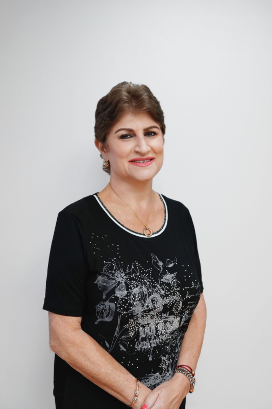 Woman with short brown hair, smiling, wearing a black shirt with floral design, standing against a white wall.