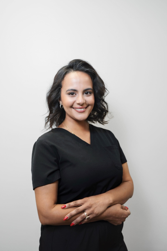 Woman with dark hair in black scrubs, arms crossed, smiling against a white backdrop.