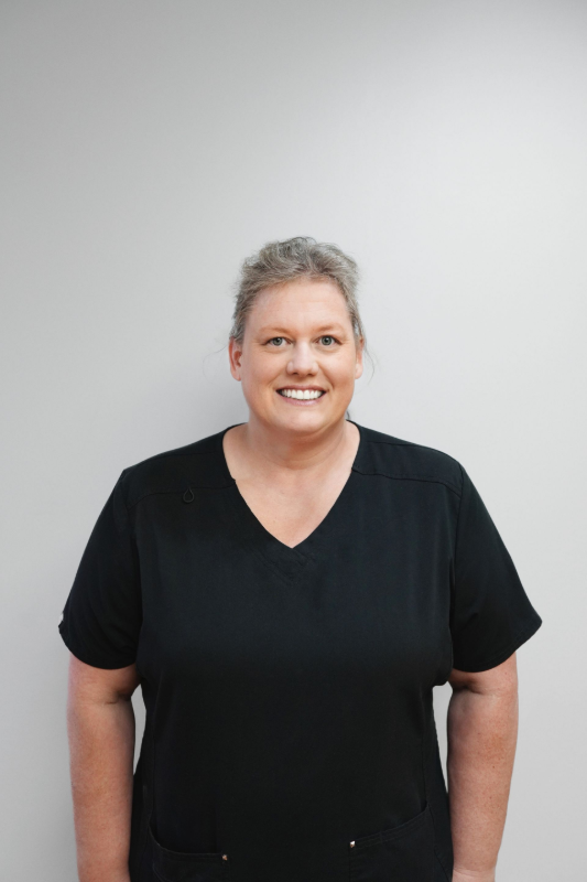 Woman with graying hair smiles, wearing a black V-neck top, standing against a plain white wall.