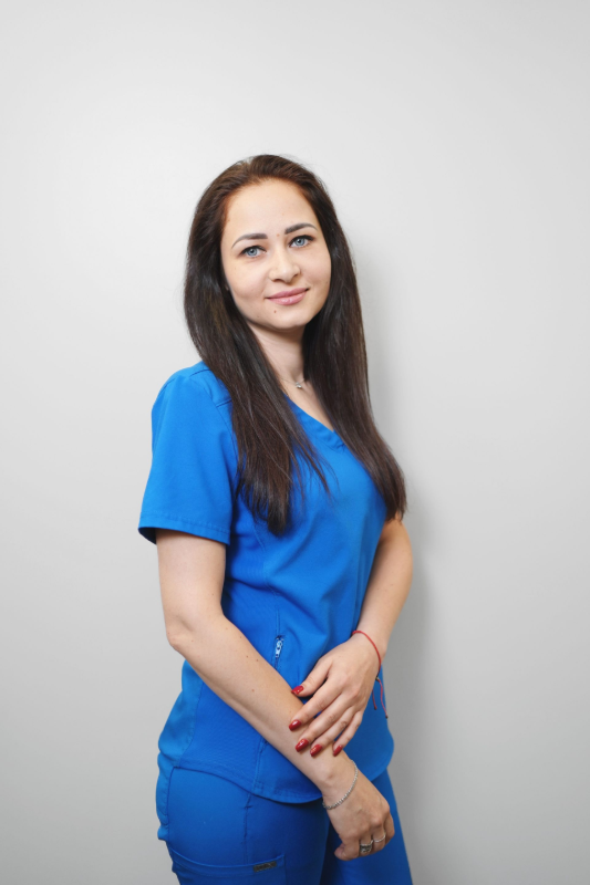 Woman in blue scrubs, smiling, standing against a plain wall.