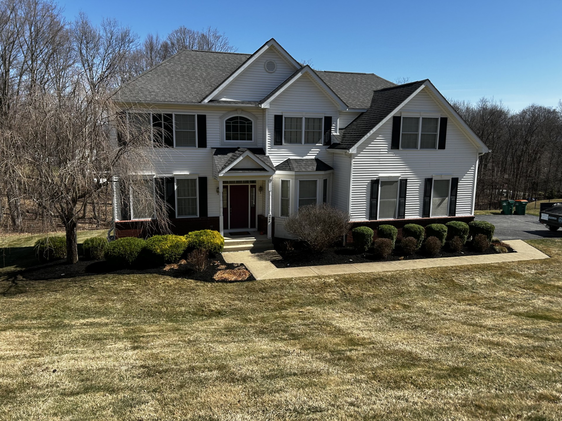 White two-story house with black shutters, red door, and landscaping, set on a grassy lawn under a blue sky.