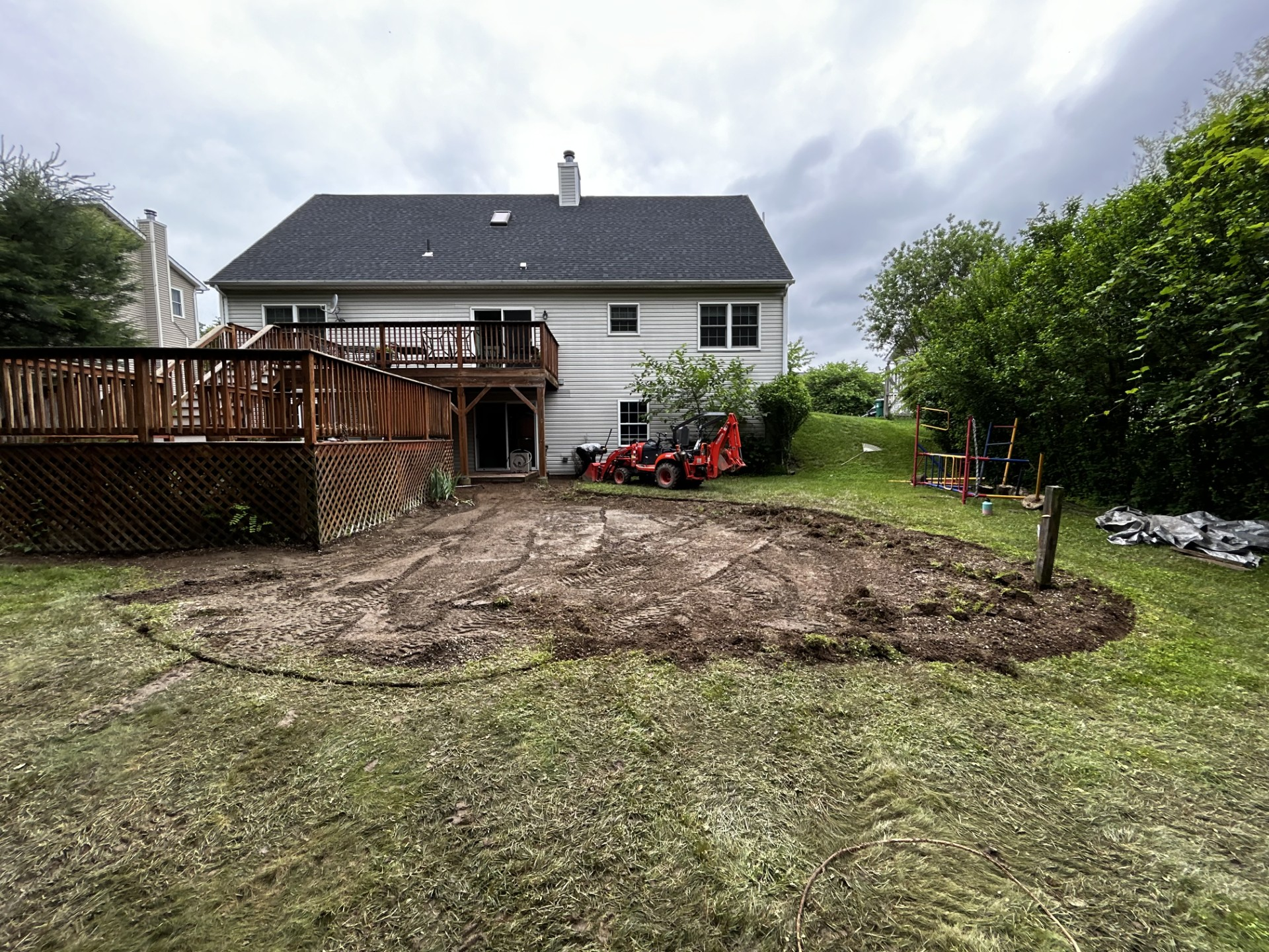 Backyard with a deck, excavated area in the center, tractor, overcast sky.