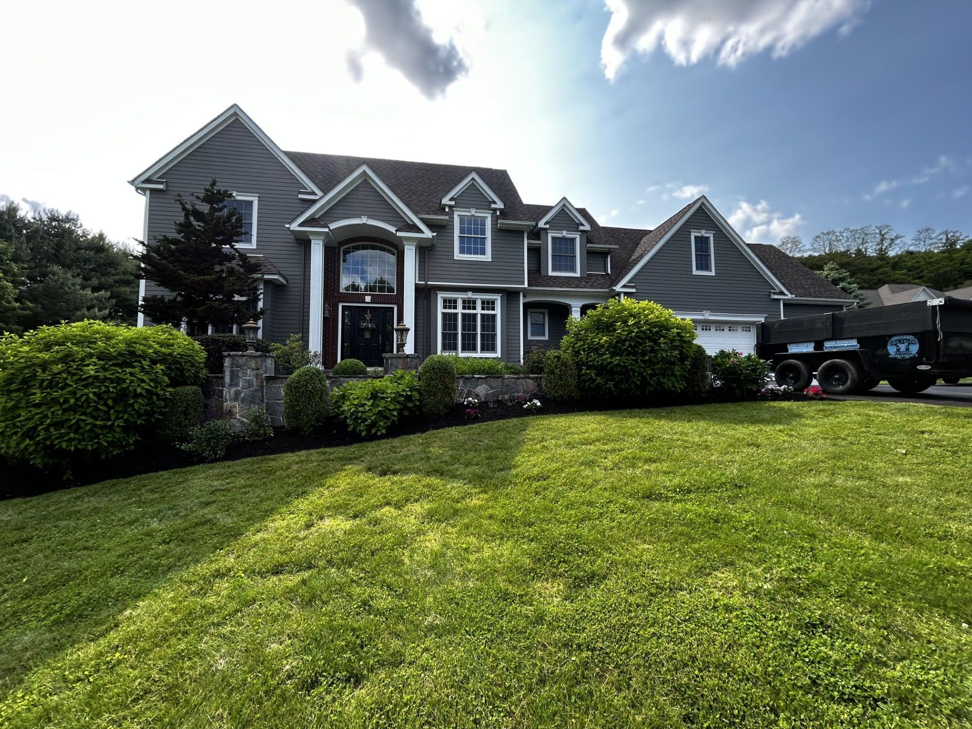 Large gray house with manicured lawn, landscaping, and a truck parked to the side on a sunny day.