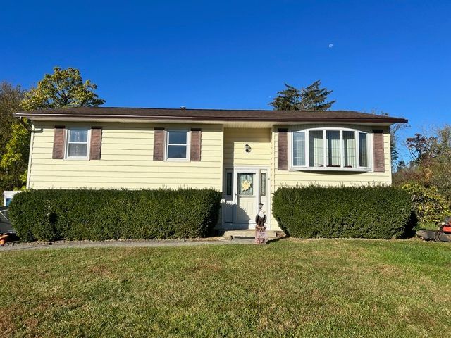 Yellow house with brown shutters, bay window, and green bushes. Blue sky with grass in front.