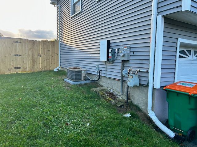 Side of a house with AC unit, electrical boxes, gas meter, and trash bin near a wooden fence and garage.