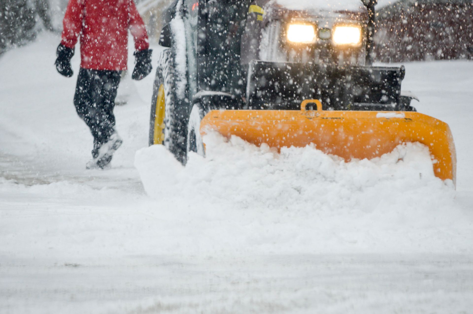 Yellow snowplow clearing snow from a driveway, person walking to the left, snowing.