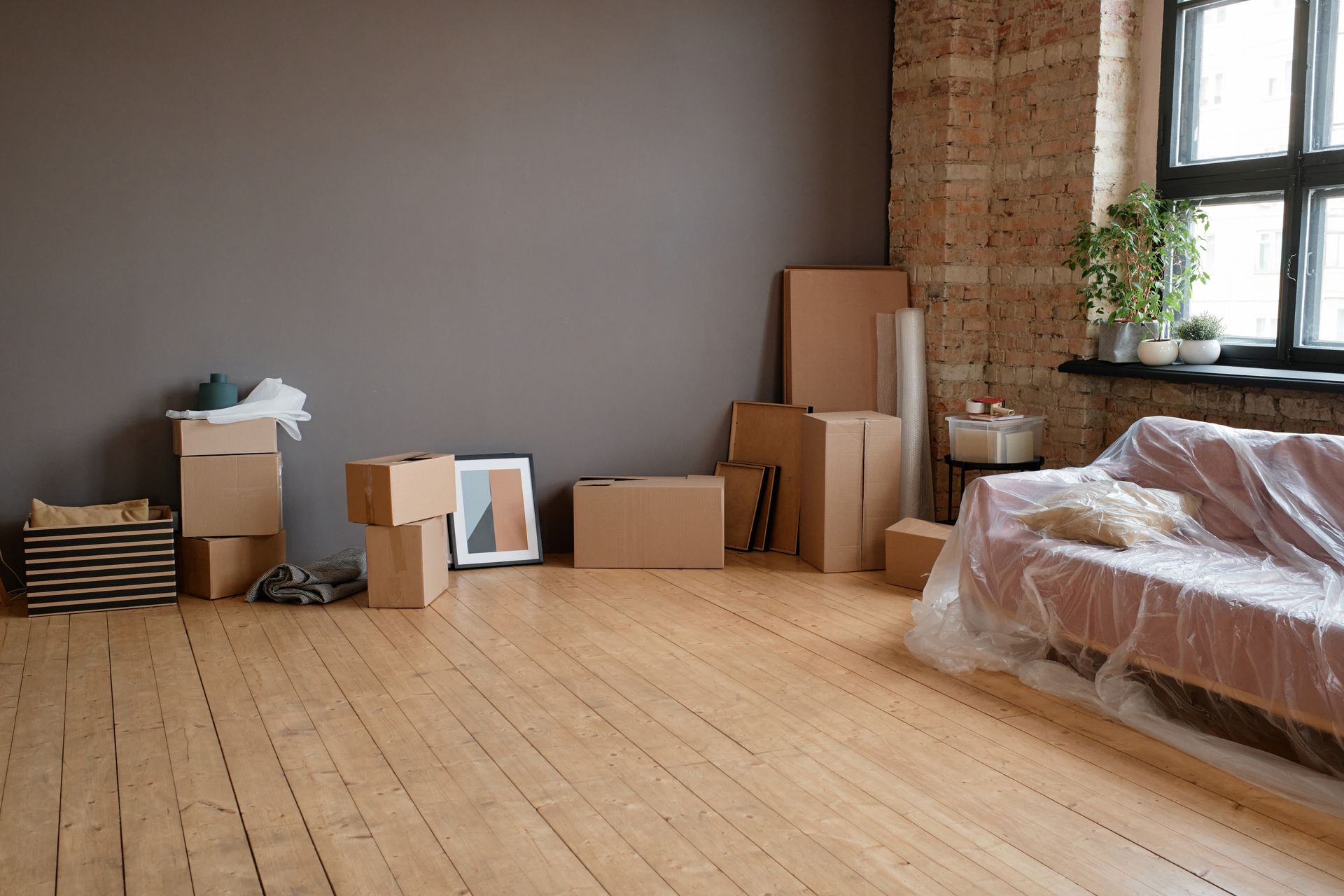 Empty room with wood floor, stacked moving boxes, and furniture wrapped in plastic near a window.