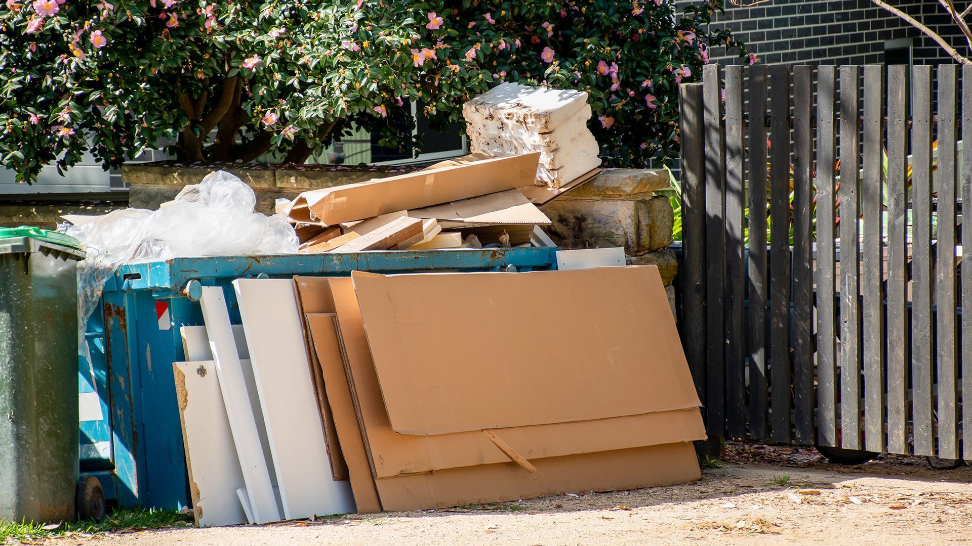 Overflowing blue recycling bin with cardboard, trash bags, and wood fence.