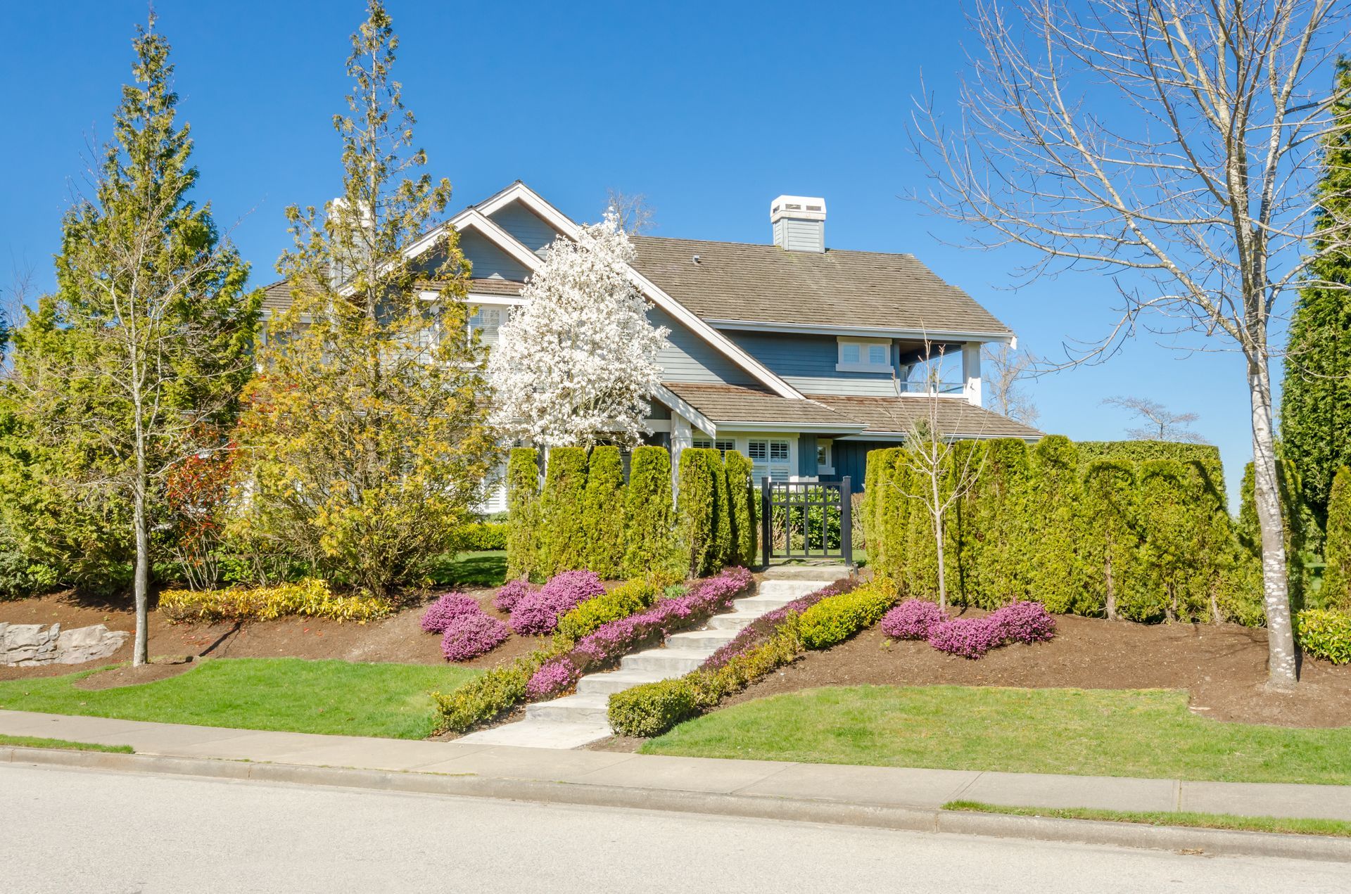 Suburban house with gray siding, white trim, manicured lawn, shrubs, and rock landscaping under a blue sky
