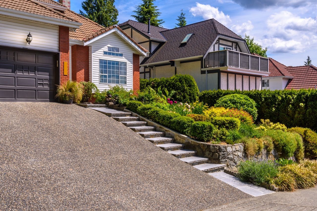 Suburban house with steep driveway, stairs, shrubs, and a tiled roof under a clear sky