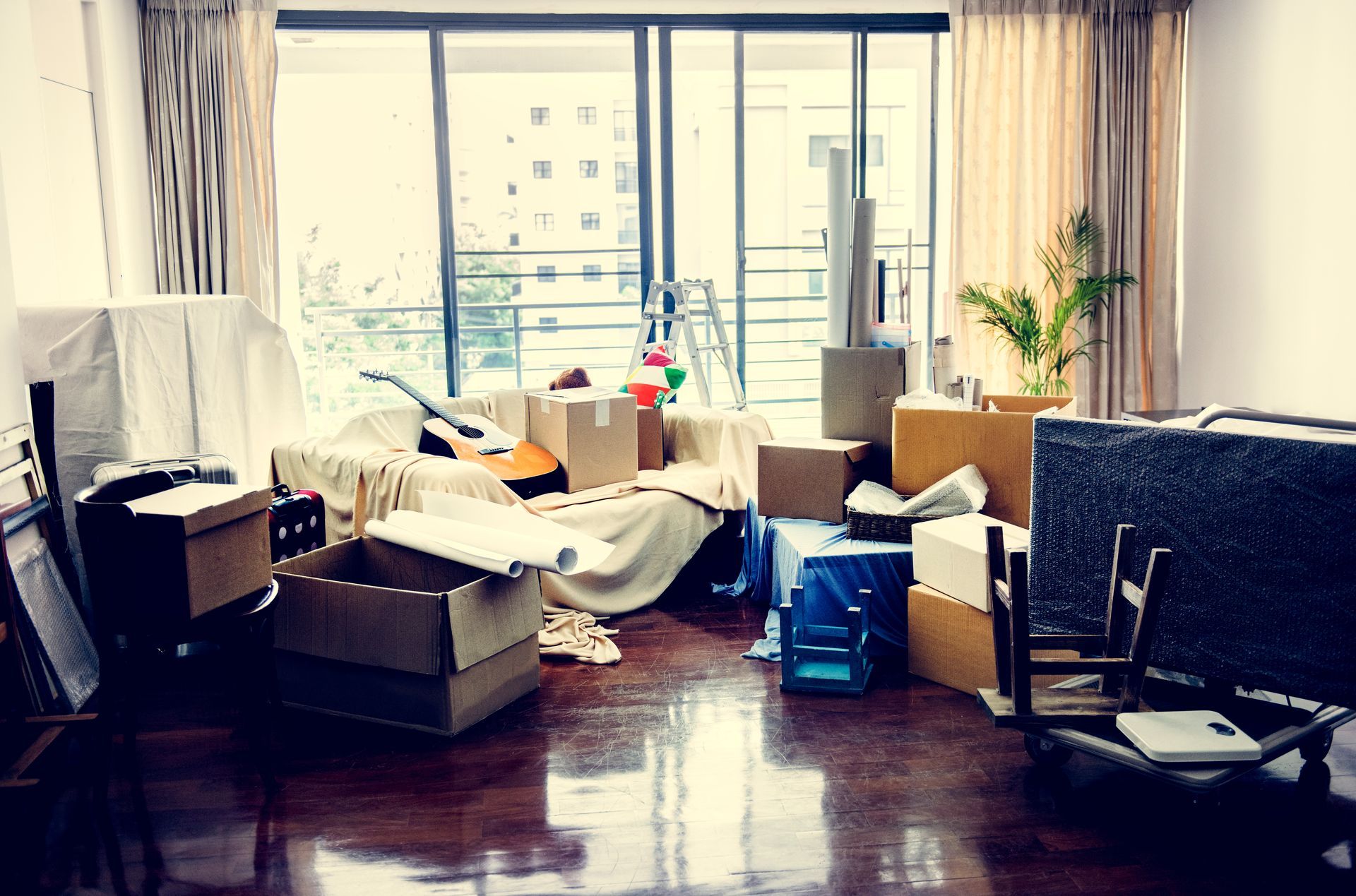 Cluttered living room with furniture, boxes, and bags in front of large windows and curtains