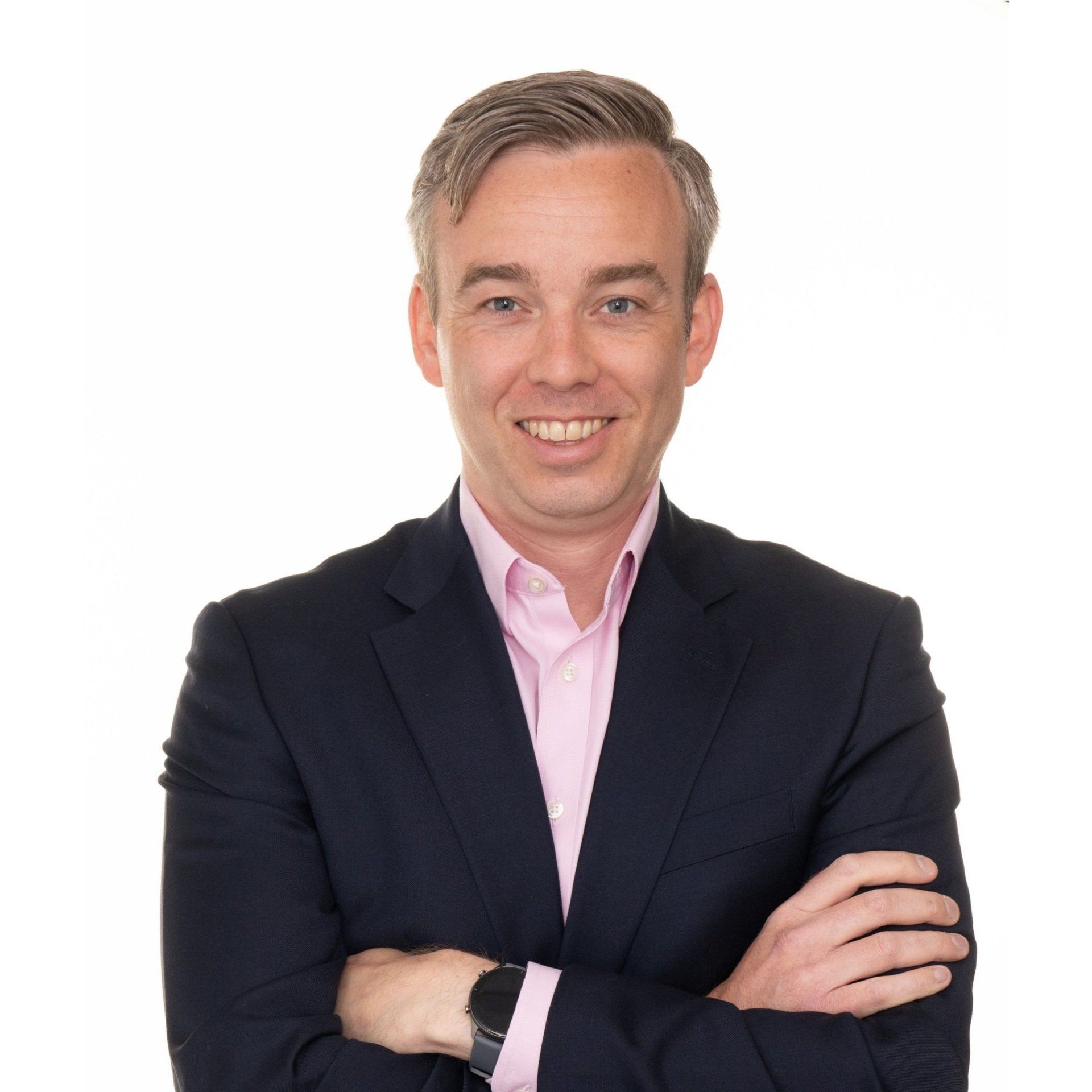 Man in a suit smiles with arms crossed, wearing a pink shirt. Studio shot with a white background.