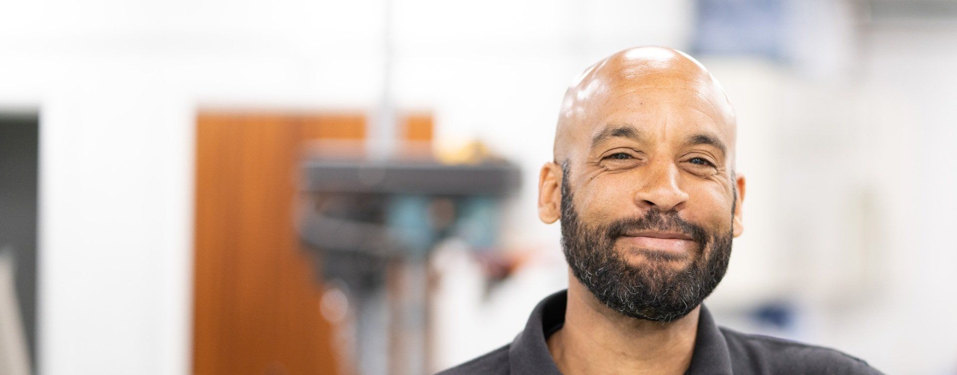 A man with a bald head and a full beard smiles at the camera, wearing a dark shirt.
