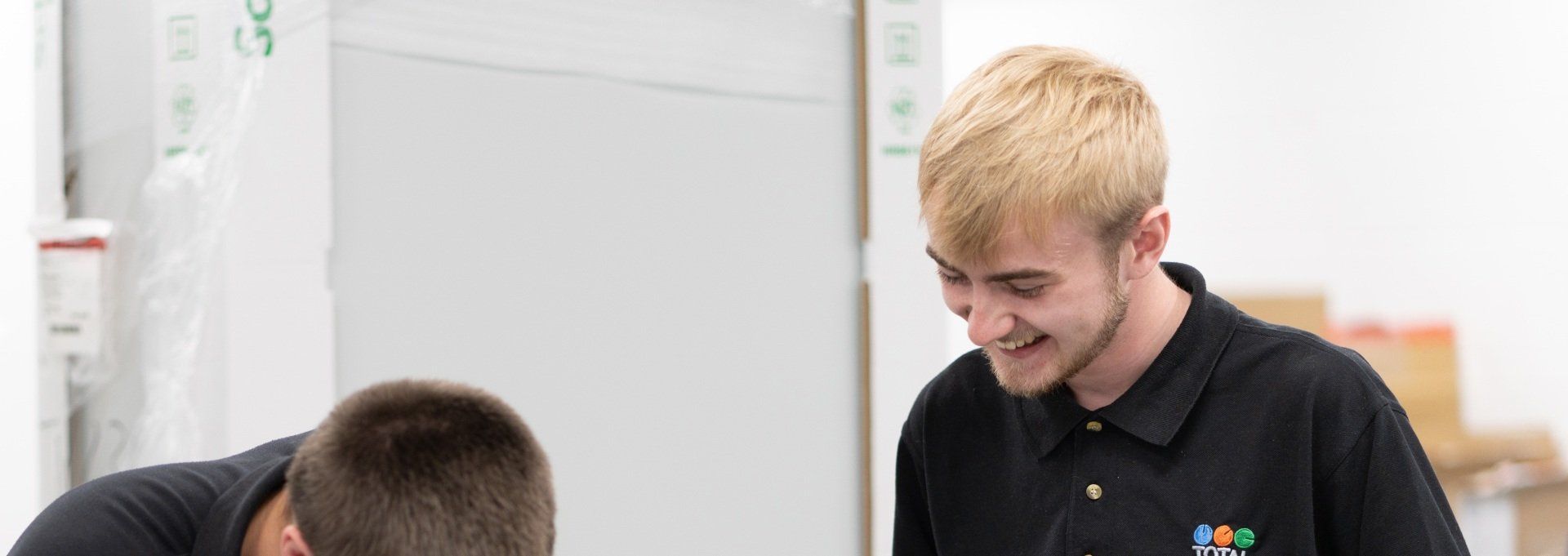 Two men smiling, one with blonde hair, in black polo shirts. Indoors, working at a table.