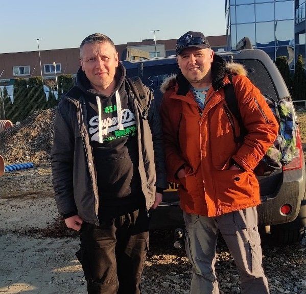 Two men standing in front of a vehicle and a building; one in an orange jacket, one in a black hoodie.
