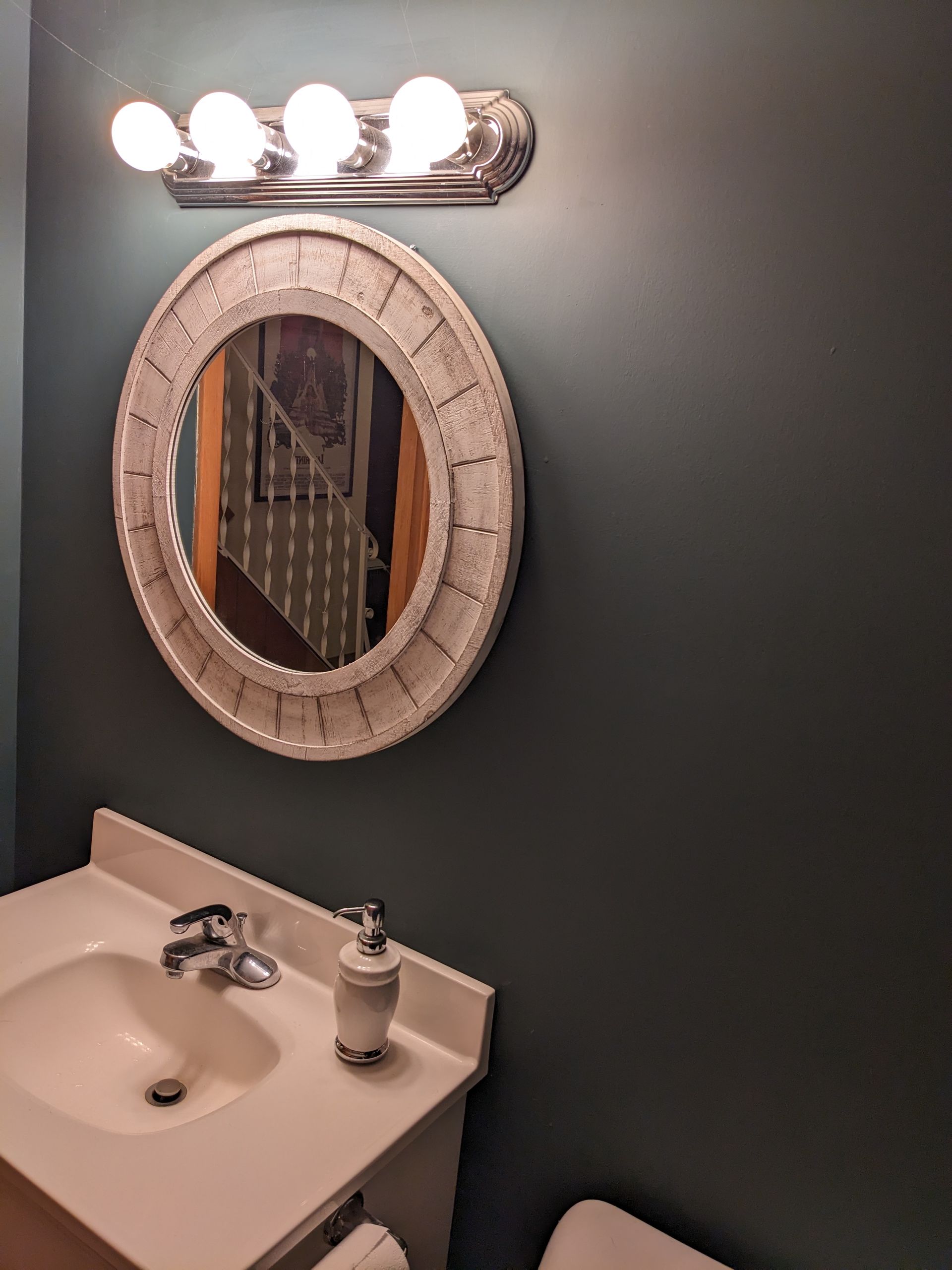 A white bathroom vanity with a basin sink and a circular, distressed white frame mirror on a dark teal wall.