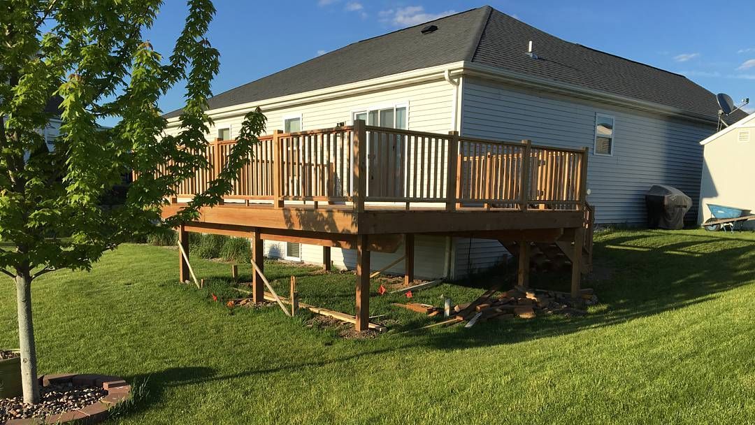 A wooden deck with railings attached to the back of a light-colored, single-story suburban house on a sunny day.
