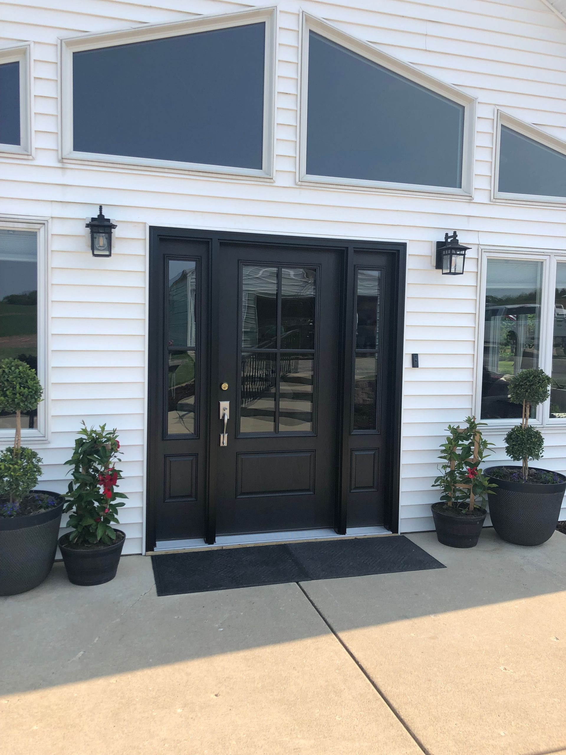 Black front door with sidelights on a white-sided building, flanked by two potted plants and matching wall lanterns.
