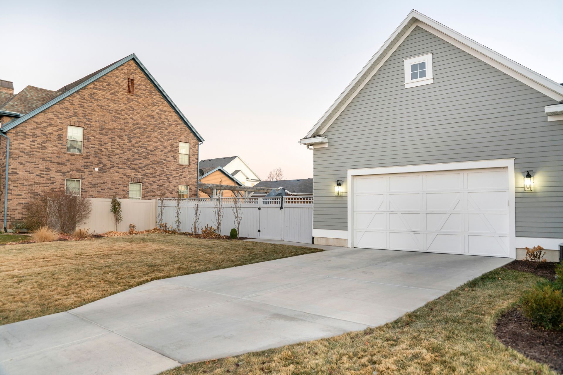 A light gray house with a white garage door stands beside a brick house with a white fence on a suburban street.