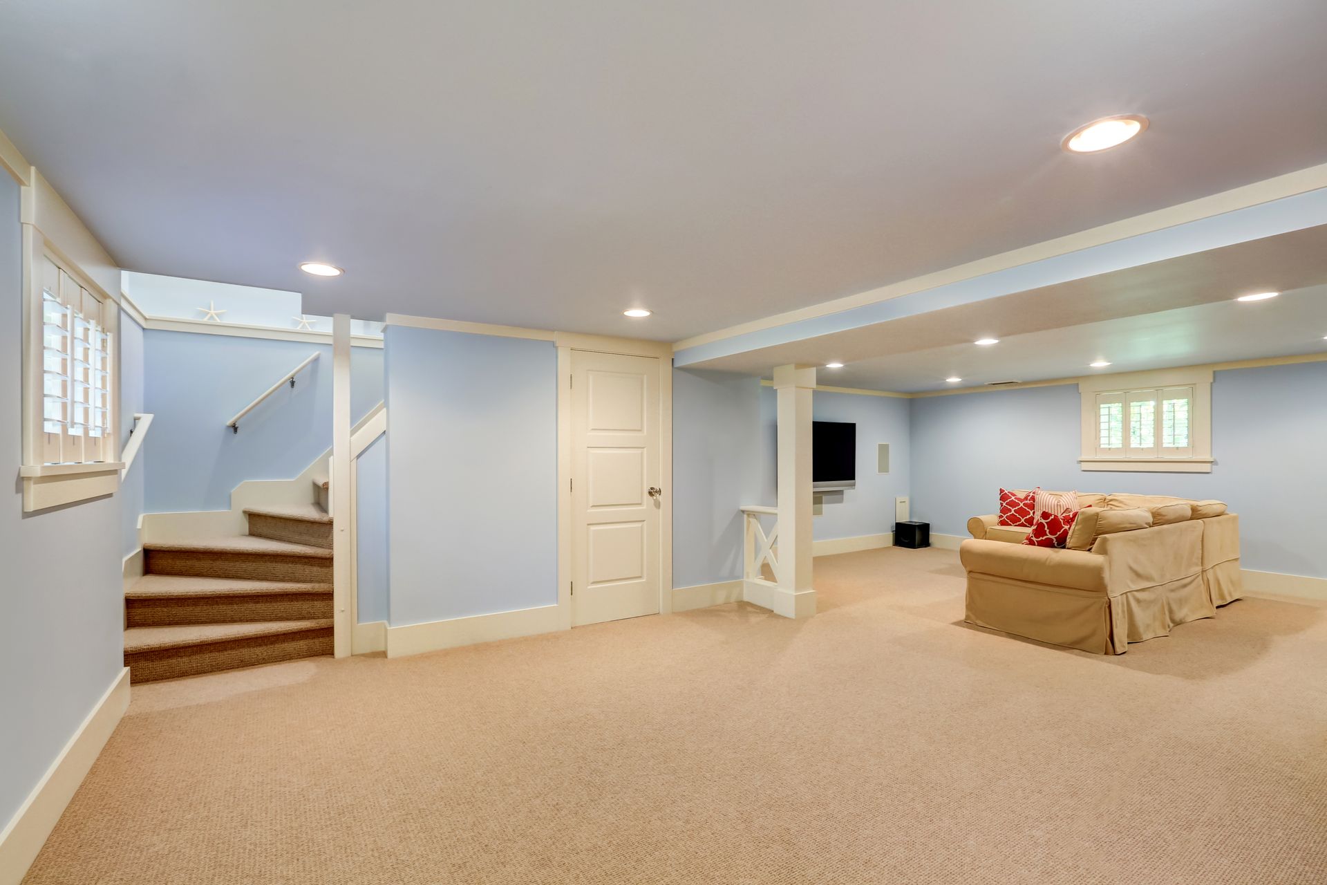 A finished basement with light blue walls, beige carpet, a wooden staircase, a closed door, and a slipcovered sofa.