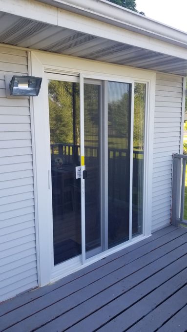 A white sliding glass door with a screen, installed in light gray vinyl siding on a wooden deck.