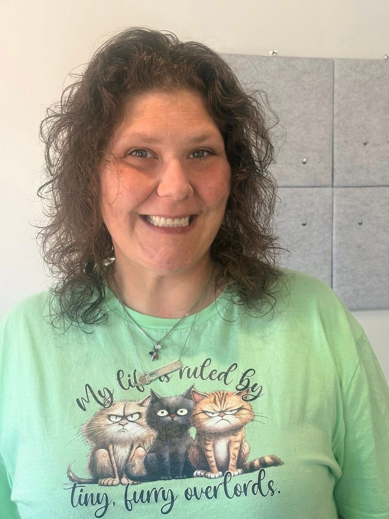 Woman in a green shirt with cat graphic smiles; medium brown curly hair; background is a gray panel.