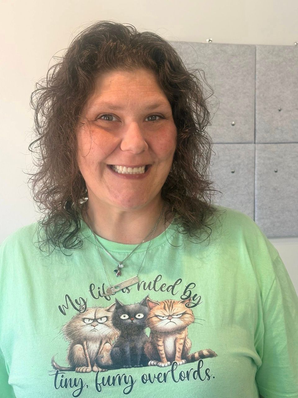 Woman in a green shirt with cat graphic smiles; medium brown curly hair; background is a gray panel.