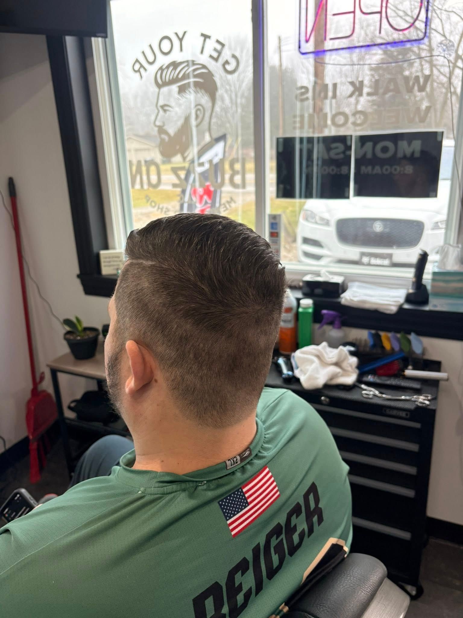 Person in barber chair getting a haircut. Barber shop interior. He is wearing a green shirt with an American flag.