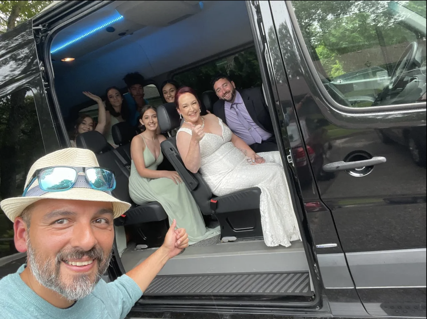 Man taking selfie with wedding party in a black van. Bride in white dress, others in formal attire.