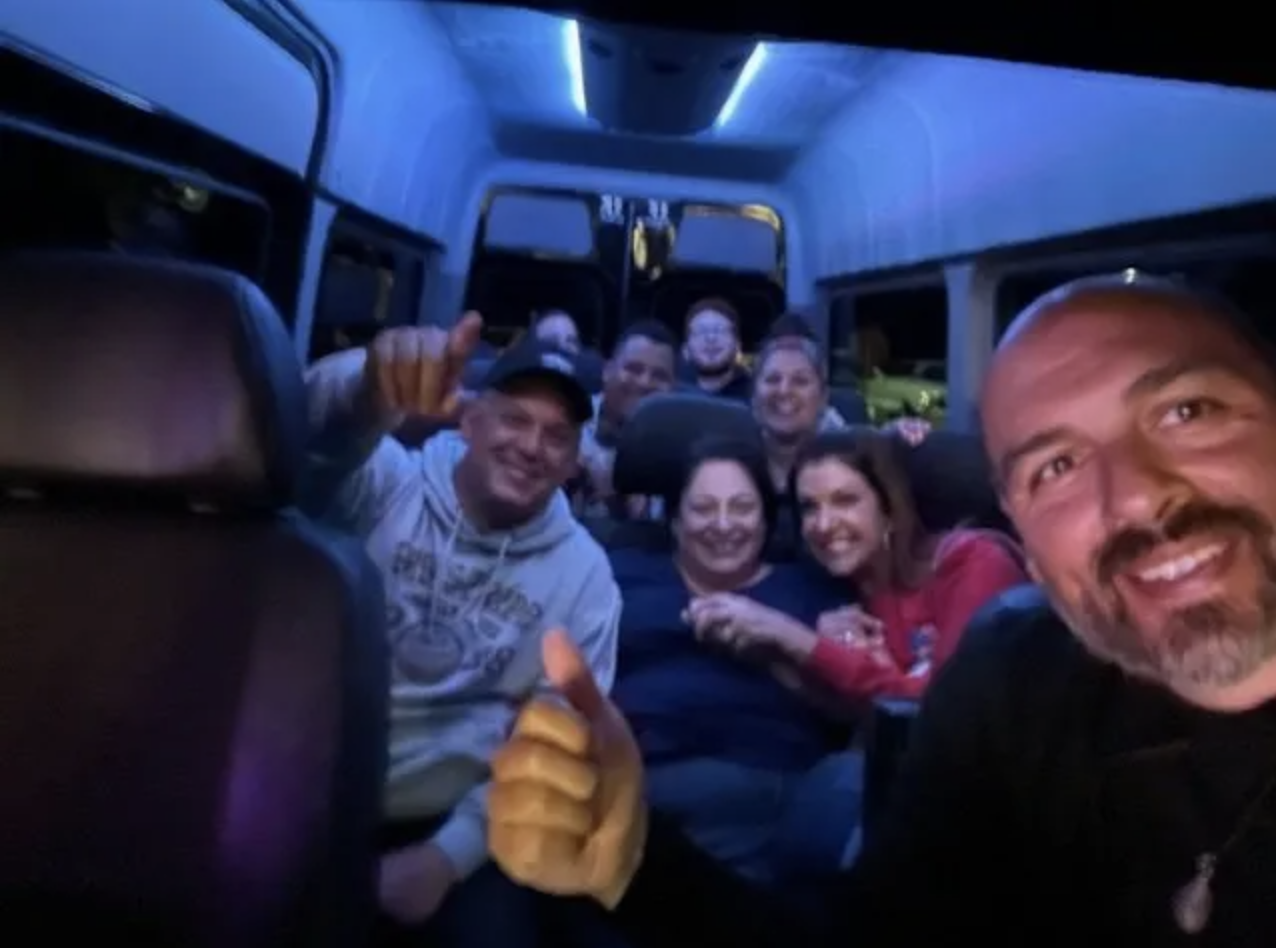 People in a van with blue interior lighting, some smiling and giving thumbs up.