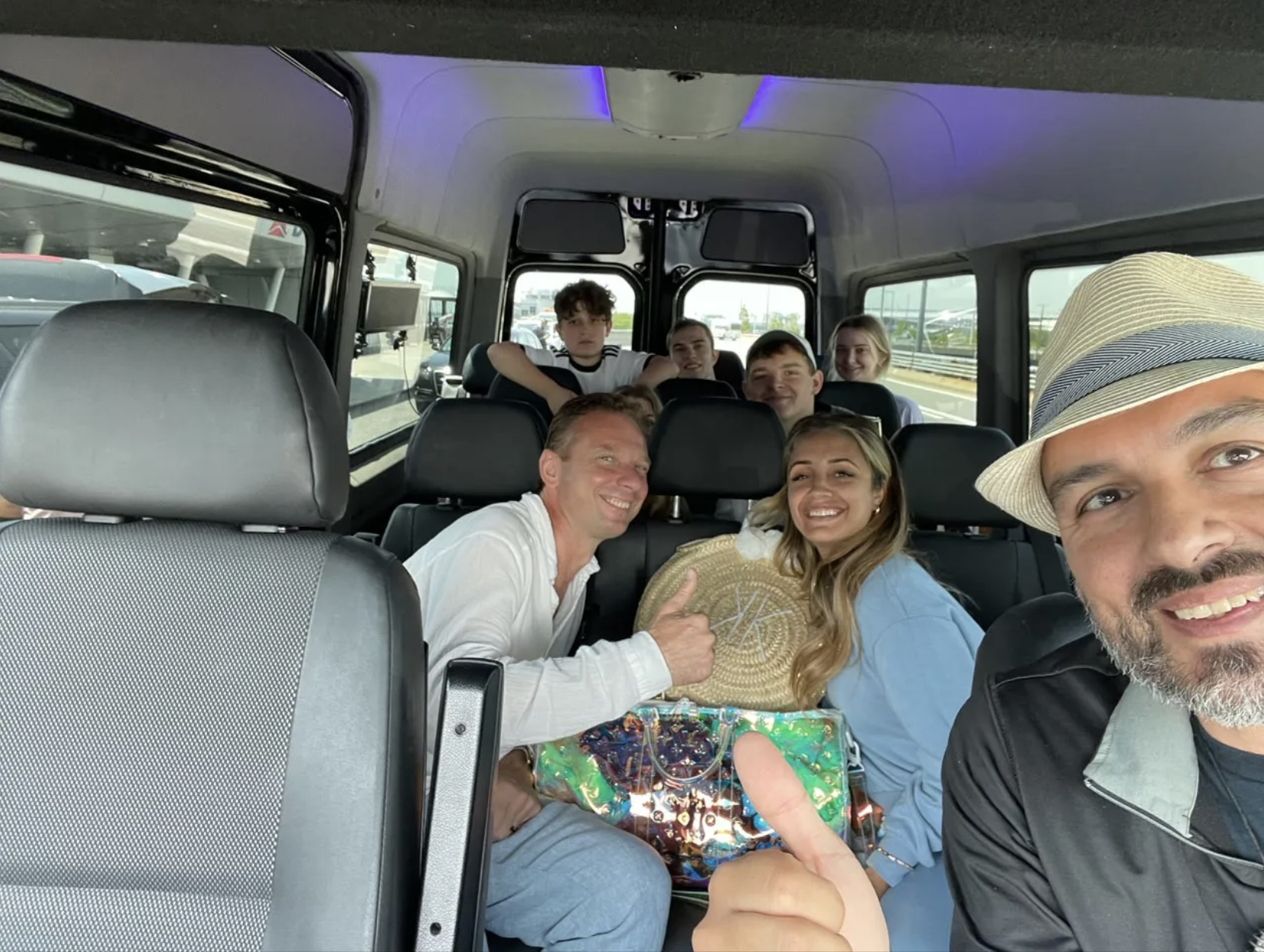 People smiling inside a van, one giving a thumbs-up. Passengers include a person in a straw hat.