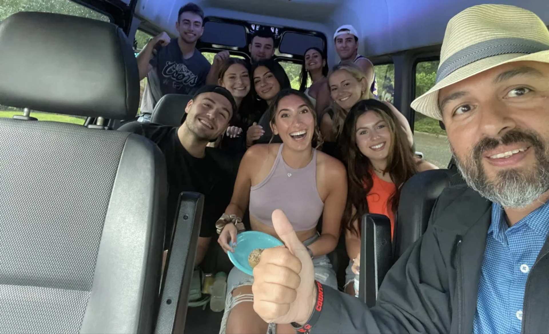 Group of people smiling in a van, one giving a thumbs up. Focus is on a smiling man in a hat taking a selfie.