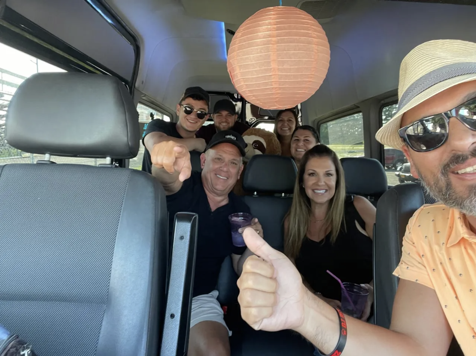 Group of people smiling in a van, one man gives thumbs up, a paper lantern hangs from the ceiling.