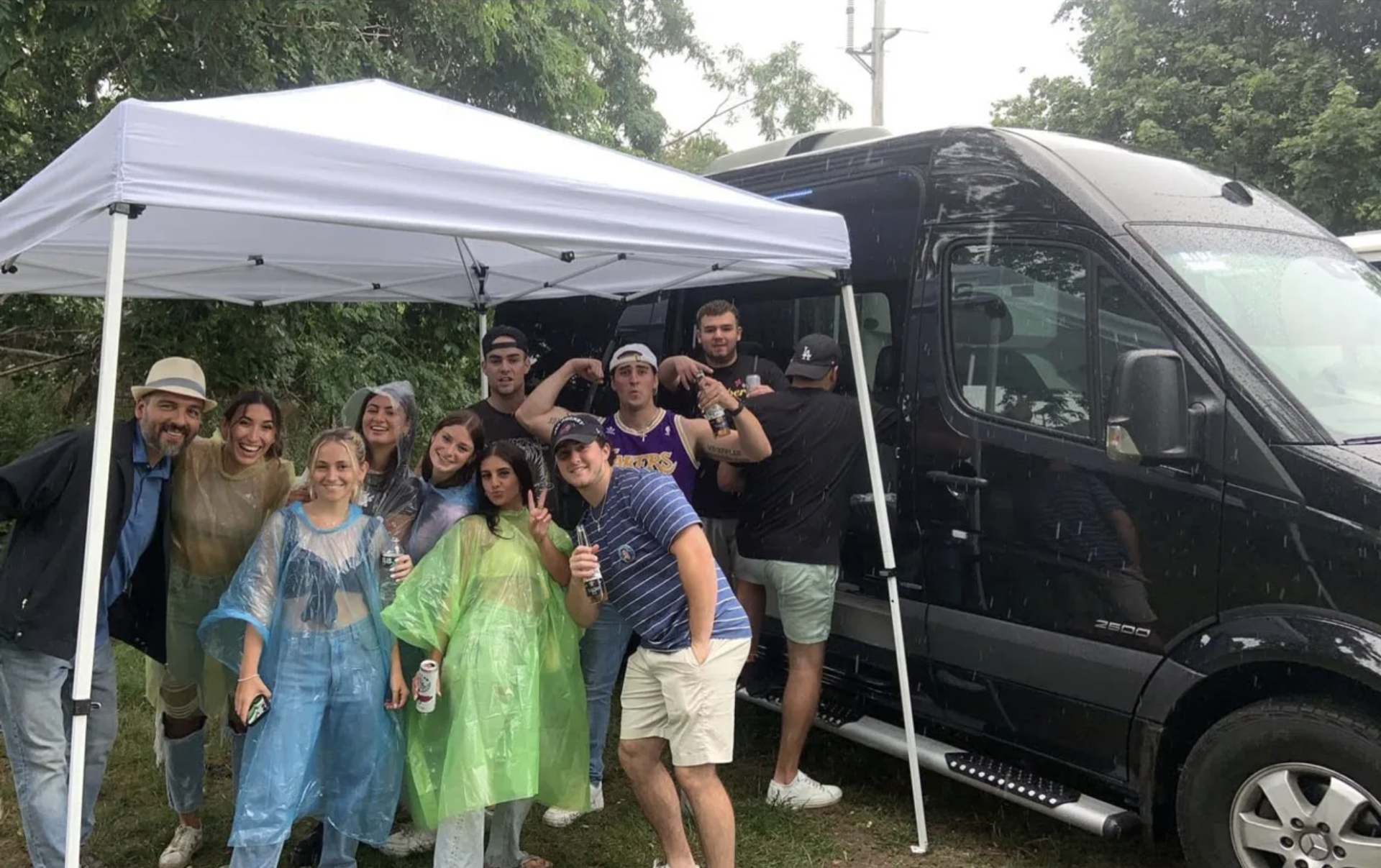 Group of people under a canopy next to a black van. Some wear rain gear and make gestures.