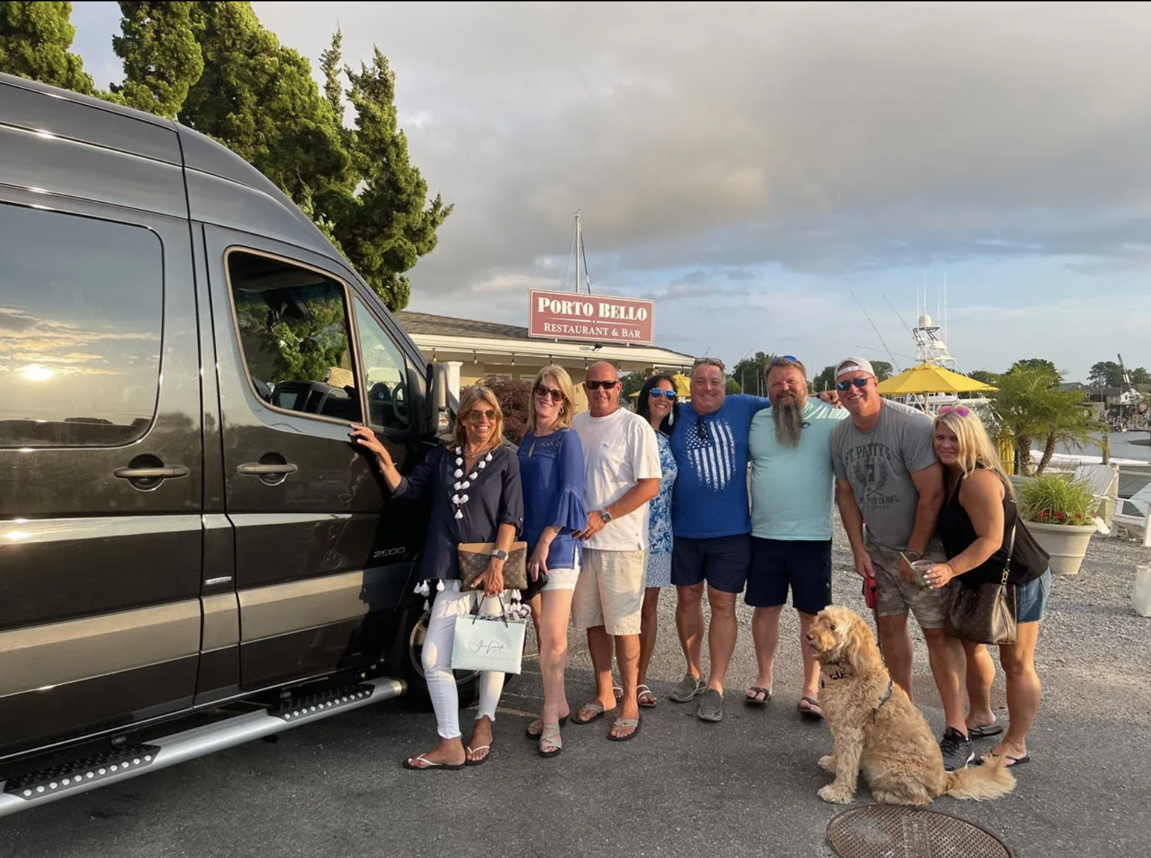 Group of people standing near a dark van at a marina; a dog sits in front. Cloudy sky, sign in background.