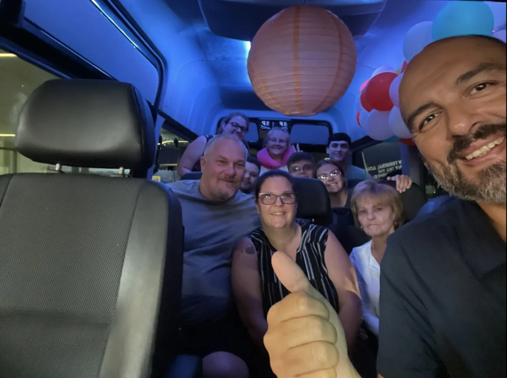 Group of people smiling inside a van, illuminated by blue lights, with balloons and a lantern. Man in front gives thumbs up.