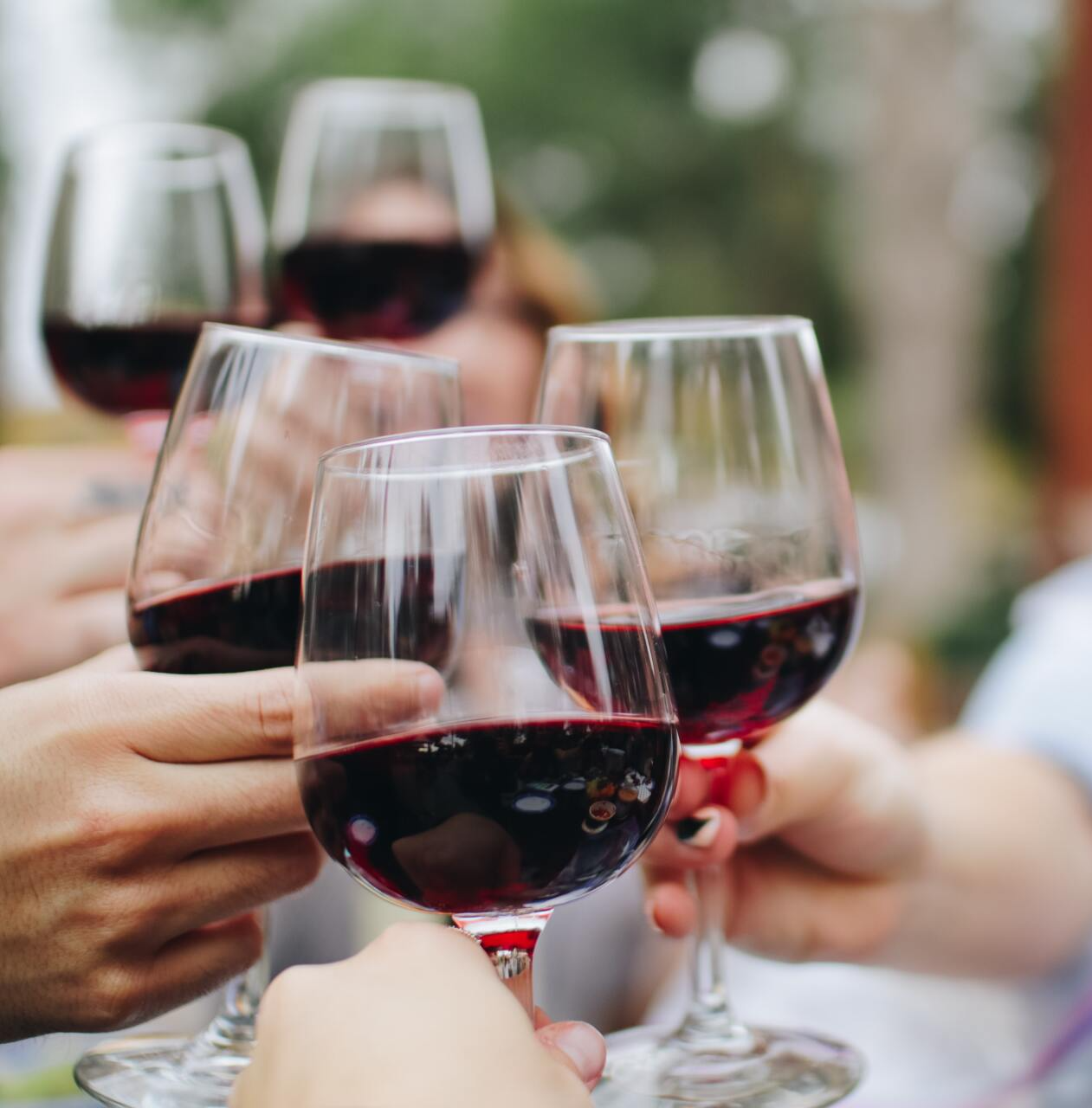 Hands clinking wine glasses filled with red wine, toasting outdoors.