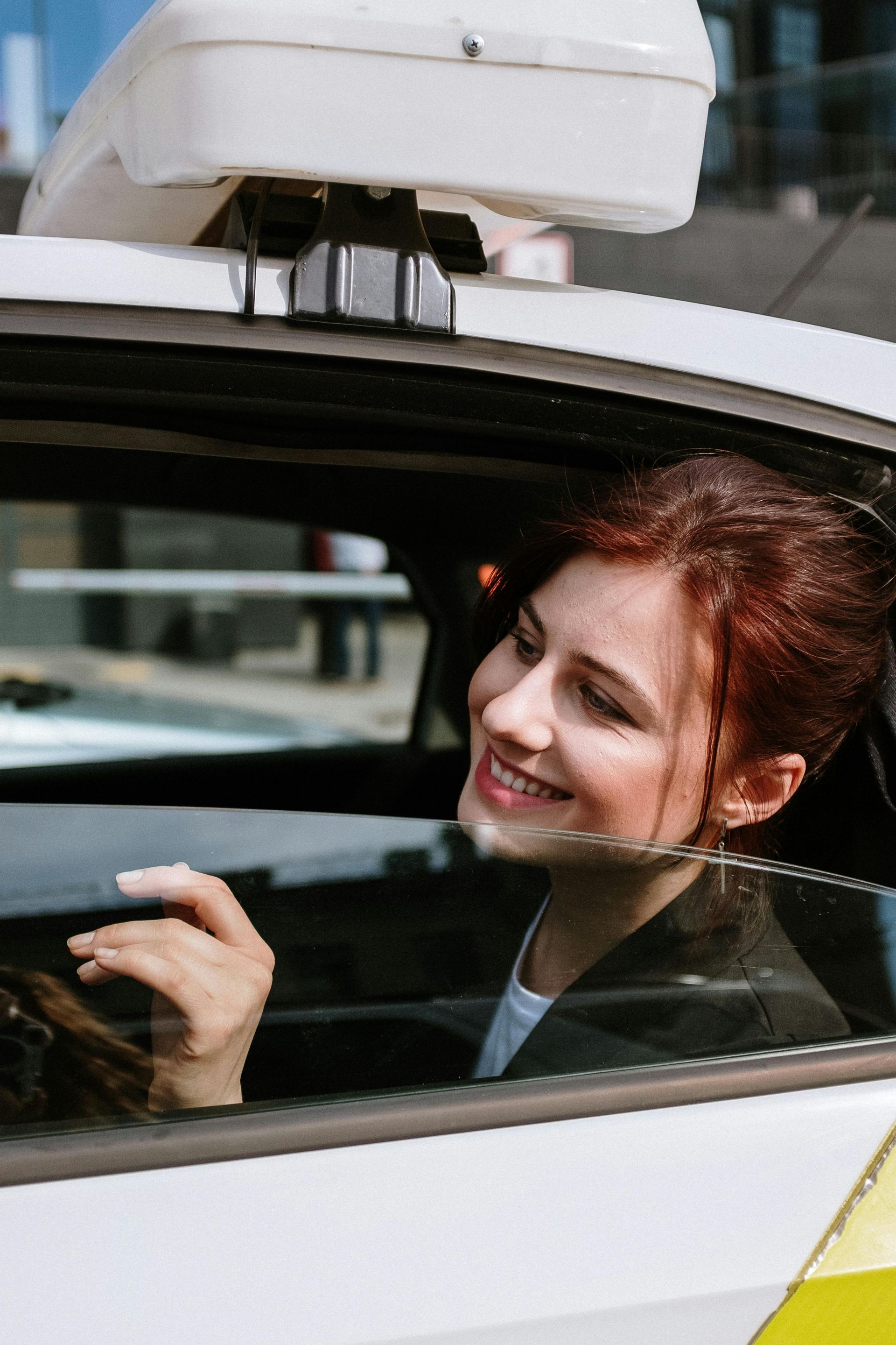 Woman in a taxi smiles, looking out the window; white taxi with sign on roof.