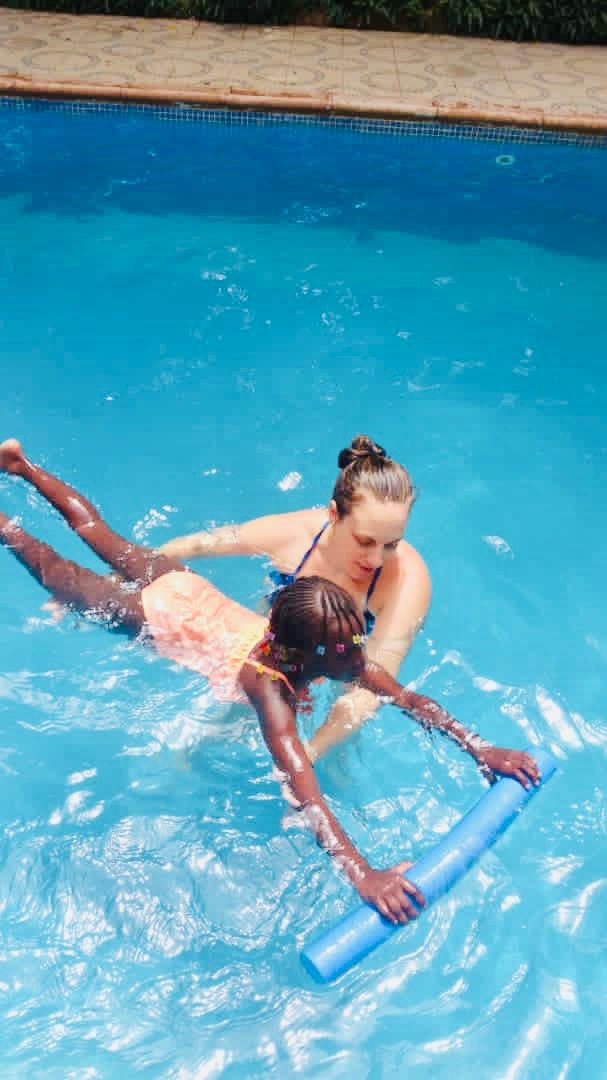 Two people in a pool using a blue float, with one person guiding the other on the water