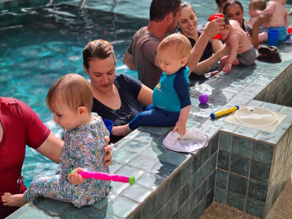 People relaxing beside a tiled pool, with children in the water holding colorful toy water guns
