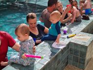 People relaxing beside a tiled pool, with children in the water holding colorful toy water guns