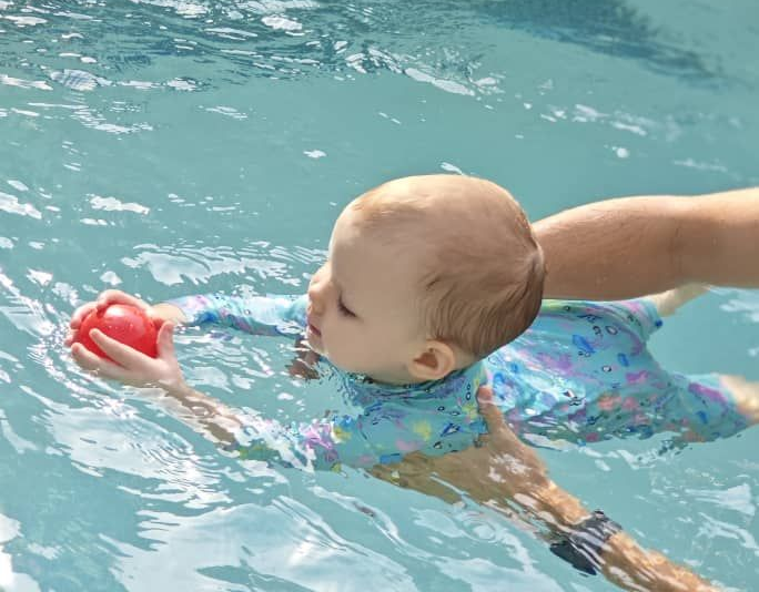 Baby swimming in a pool, reaching for a red toy while held by an adult arm