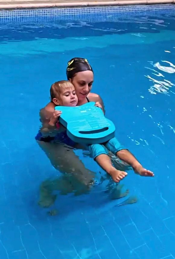 Woman holding a child in a blue swimming pool, both wearing blue flotation devices