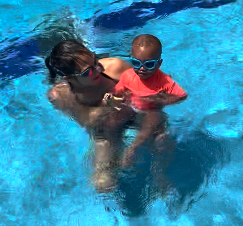 Adult holding a child in a bright blue swimming pool, both wearing goggles