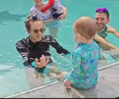 People in a swimming pool, with one person holding a child at the edge and others smiling nearby