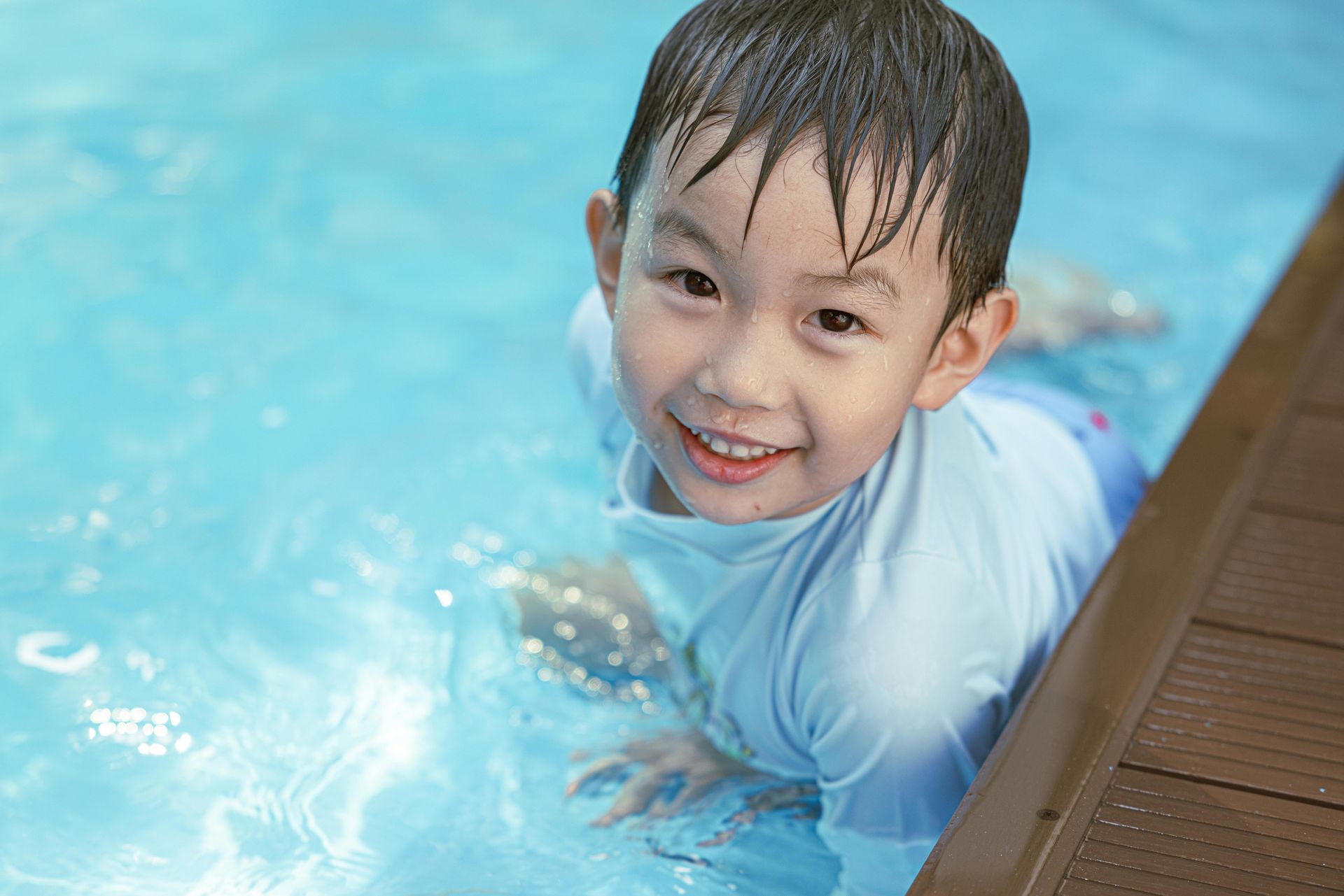 Smiling child in a light blue shirt playing in a swimming pool beside a wooden deck