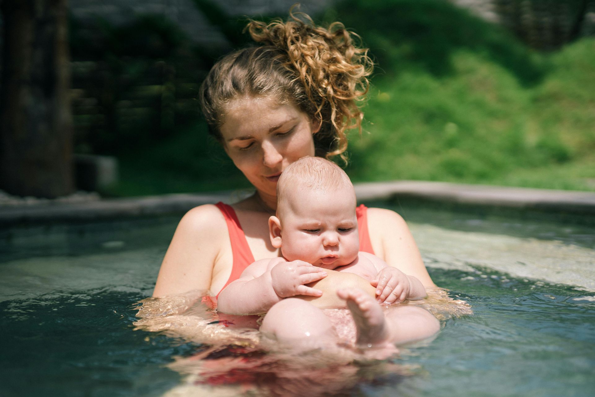 Adult holding a baby in a pool, both in water near the edge with green foliage behind them