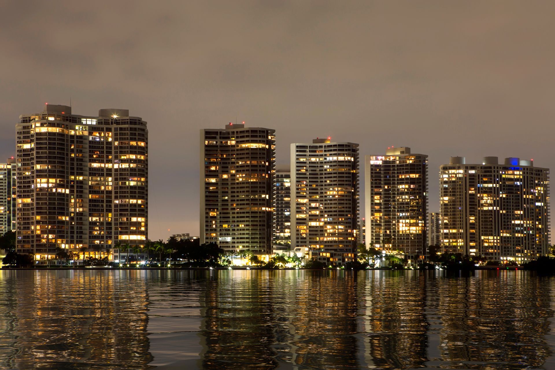 Skyscrapers with lit windows reflect in calm water at night.