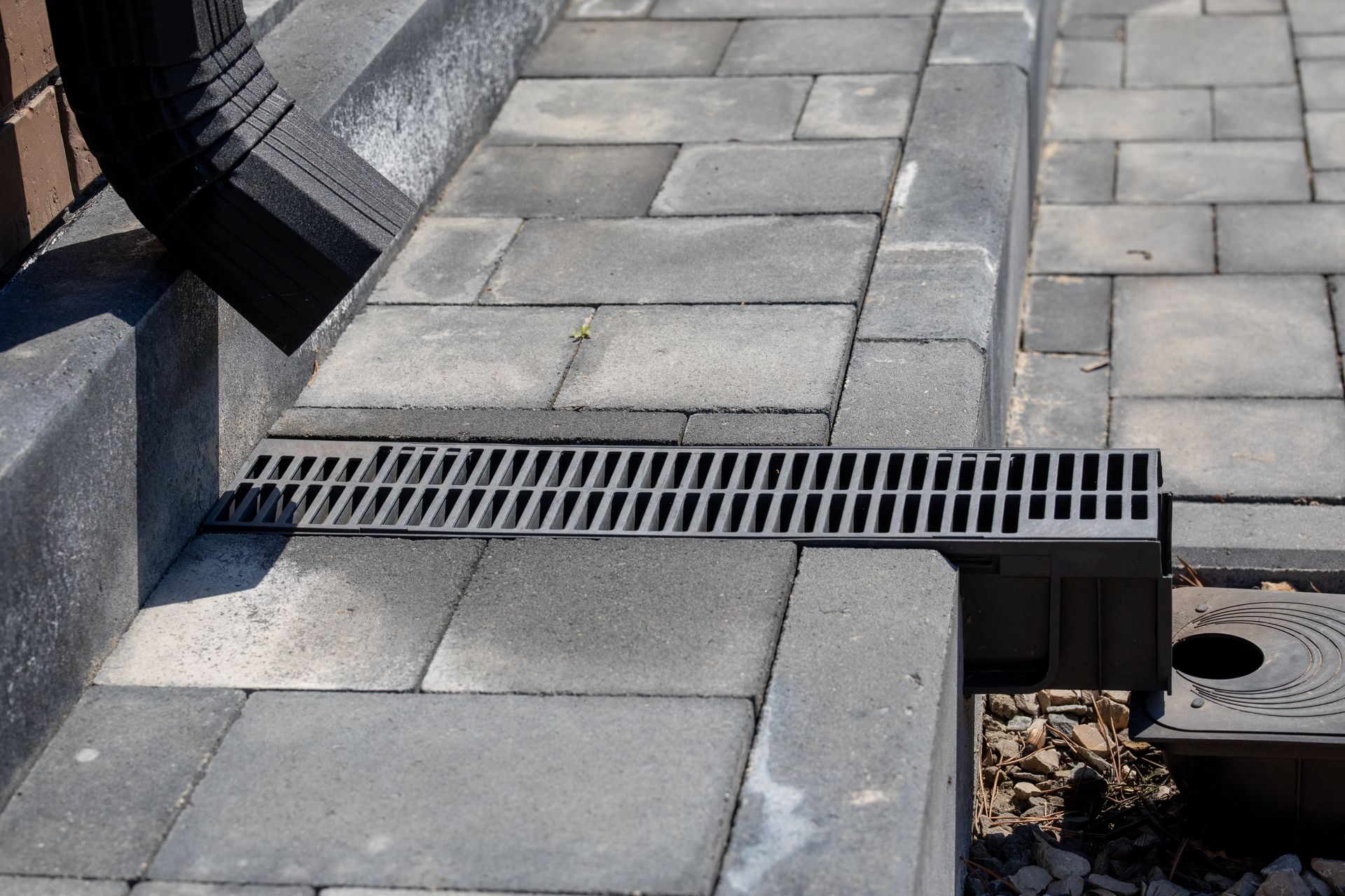 Sidewalk paving stones with a metal drainage grate beside a curb and building wall