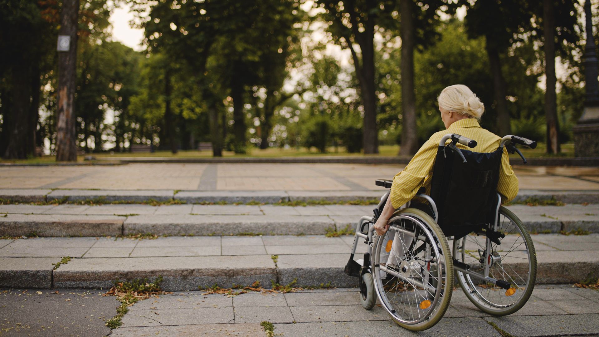 An elderly woman in a wheelchair is sitting on a sidewalk with steps.